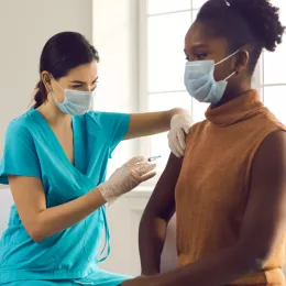 healthcare worker in blue scrubs giving a woman a vaccine shot in her upper arm in front of a bright window